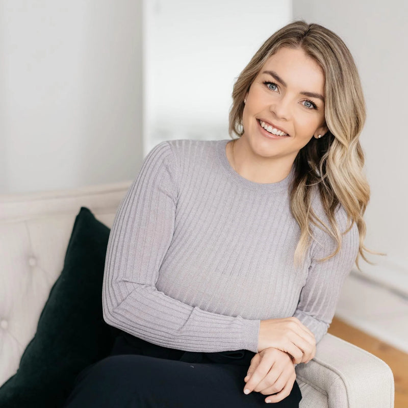 Tara O'Kelly, foiunder of TOK, sitting on a couch wearing a gray sweater in a home setting